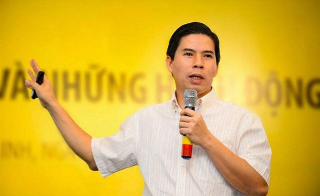 Người đàn ông cầm micro phát biểu trước phông nền màu vàng, mặc áo sơ mi sáng màu.