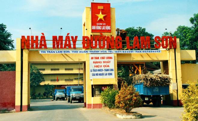 Cổng vào nhà máy đường với xe tải chở mía, biển hiệu lớn và cây xanh xung quanh.