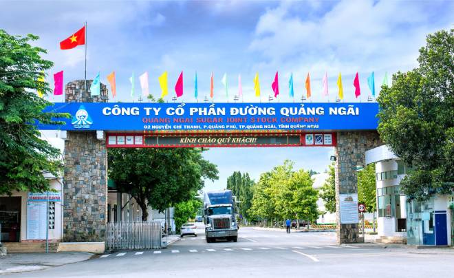 Cổng vào của một công ty với xe tải đi qua, cây xanh và cờ nhiều màu sắc phía trên.