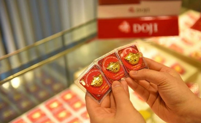 Hai tay cầm ba miếng vàng trong bao bì đỏ, phía sau là quầy trưng bày trong cửa hàng trang sức.