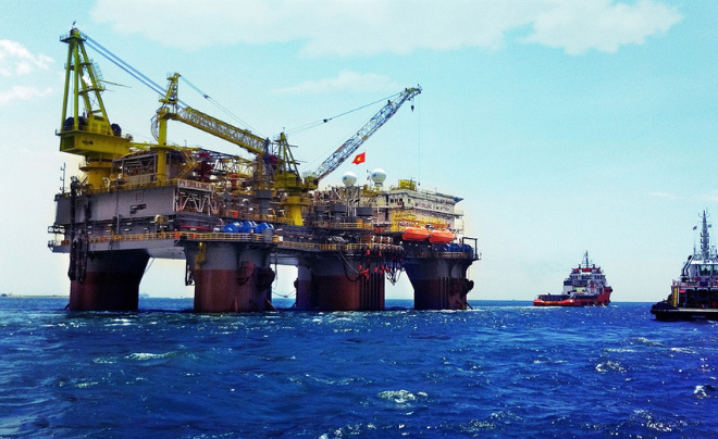 Giàn khoan dầu ngoài khơi với cần cẩu và tàu hỗ trợ trên biển xanh, cờ bay trên giàn.