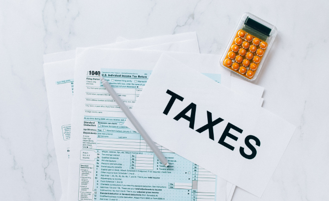 Các biểu mẫu thuế, máy tính và tờ giấy ghi "TAXES" trên mặt bàn màu trắng.