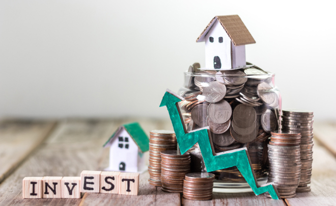 Lọ thủy tinh đựng tiền xu, mô hình nhà và chữ "INVEST" trên bàn gỗ, biểu tượng mũi tên xanh hướng lên.