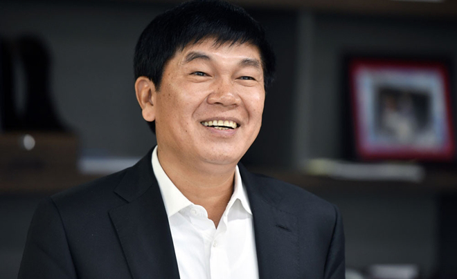 Một người đàn ông mặc vest đen và áo sơ mi trắng đang mỉm cười, đứng trước phông nền với kệ sách.