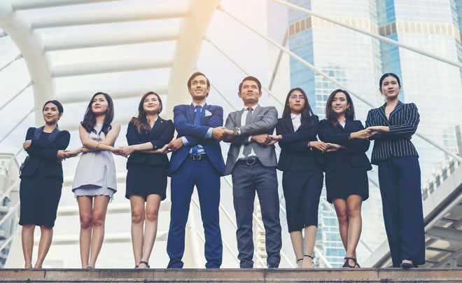 Nhóm người mặc trang phục công sở đứng xếp hàng, tay đan chéo, nền là kiến trúc đô thị hiện đại.
