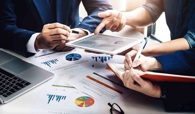 Nhóm người thảo luận tại bàn với tài liệu, máy tính xách tay và máy tính bảng. Họ xem biểu đồ và ghi chú.