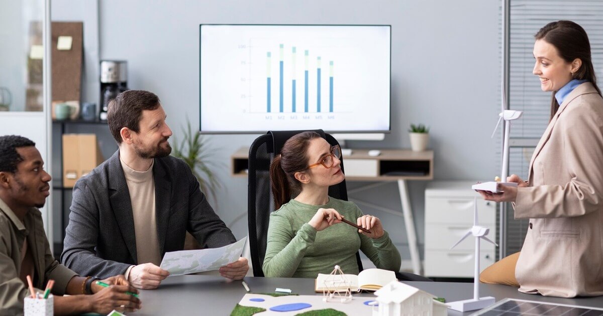 Một nhóm người đang thảo luận trong văn phòng, phía sau là màn hình hiển thị biểu đồ cột.