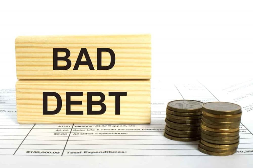 Hai khối gỗ có chữ "BAD DEBT" cạnh cột tiền xu trên nền tài liệu tài chính.