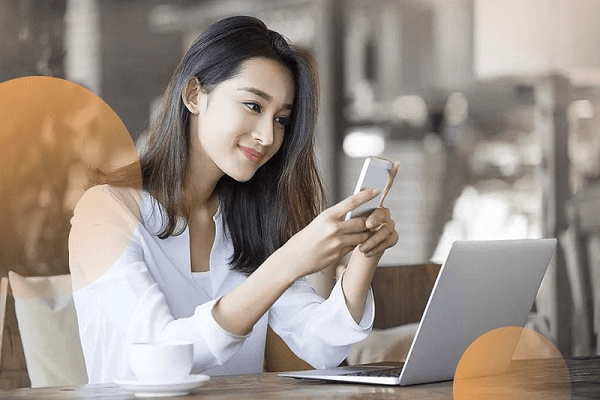 Người phụ nữ ngồi tại quán cà phê, sử dụng điện thoại và máy tính xách tay, với ly cà phê bên cạnh.