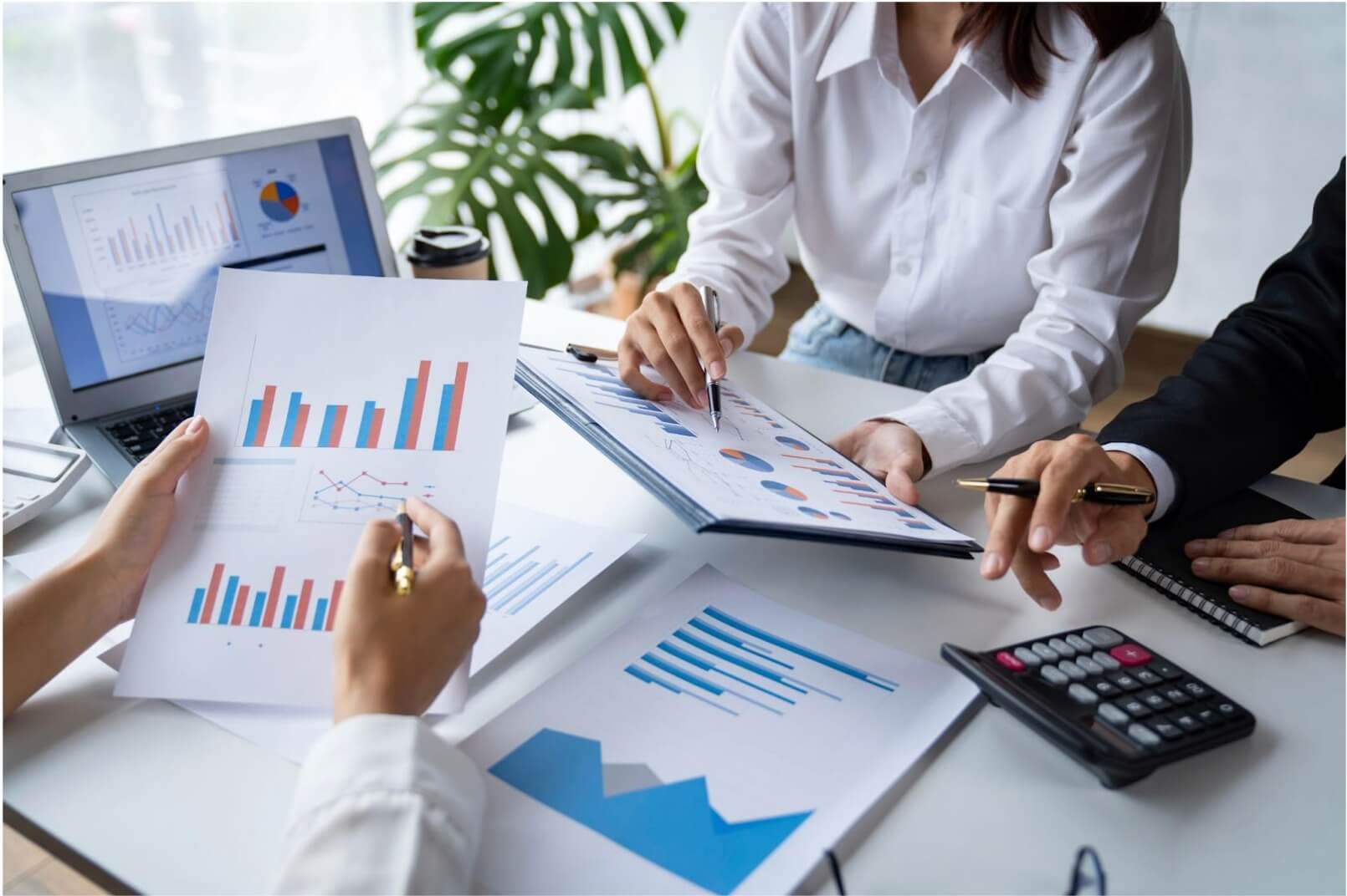 Các tay cầm tài liệu biểu đồ và bút, thảo luận xung quanh bàn với máy tính xách tay và máy tính cầm tay.