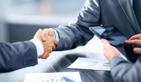 Hai người bắt tay trên bàn làm việc, bên cạnh là máy tính bảng và tài liệu.