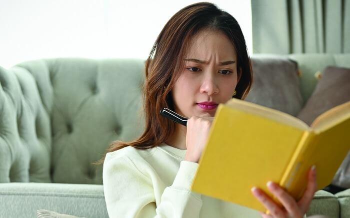 Người phụ nữ ngồi trên ghế sofa, chăm chú đọc một cuốn sách màu vàng, tay cầm bút, biểu cảm tập trung.