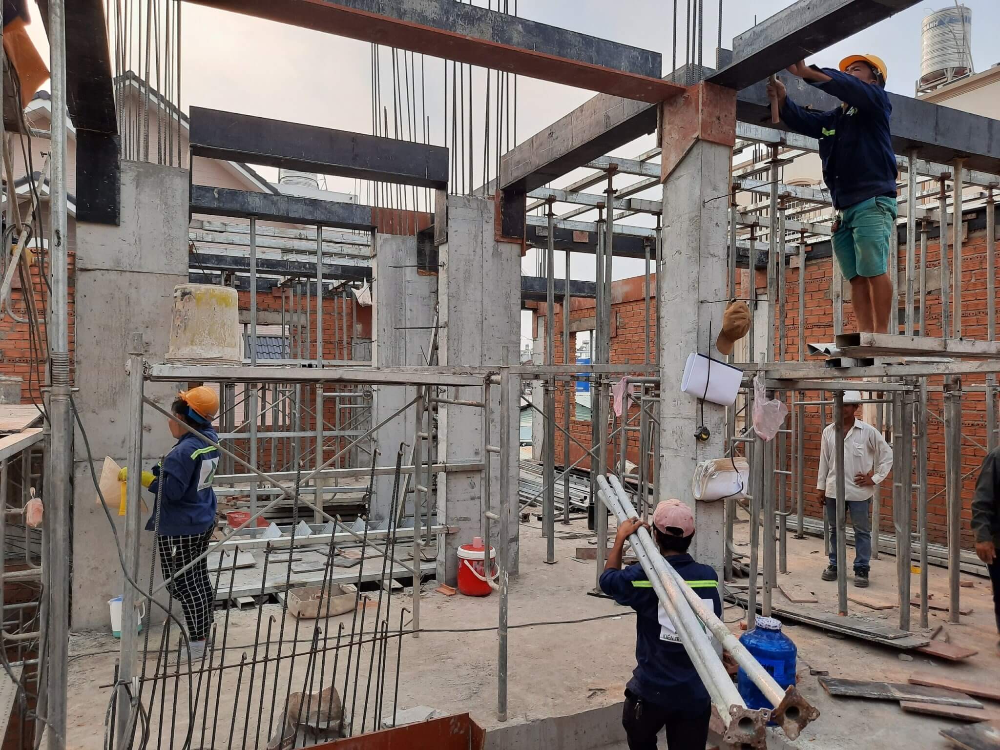 Công nhân xây dựng làm việc trên công trường với giàn giáo và cột bê tông, mang mũ bảo hộ.