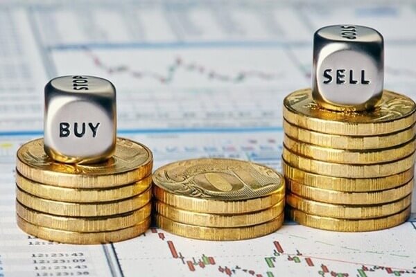 Các đồng xu xếp chồng với hai viên xúc xắc có chữ "BUY" và "SELL" trên nền biểu đồ tài chính.