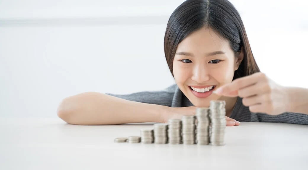 Người phụ nữ mỉm cười khi xếp chồng tiền xu trên bàn, biểu thị sự tiết kiệm hoặc đầu tư.