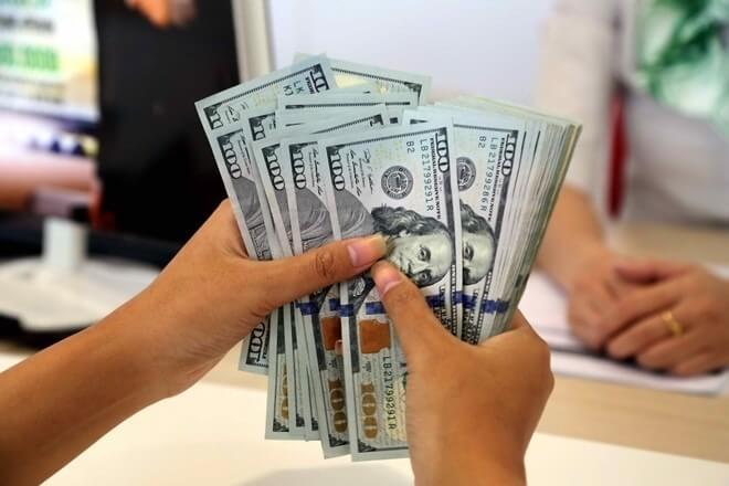 Hai tay cầm nhiều tờ tiền đô la Mỹ, một người khác ngồi phía trước trên nền bàn làm việc.
