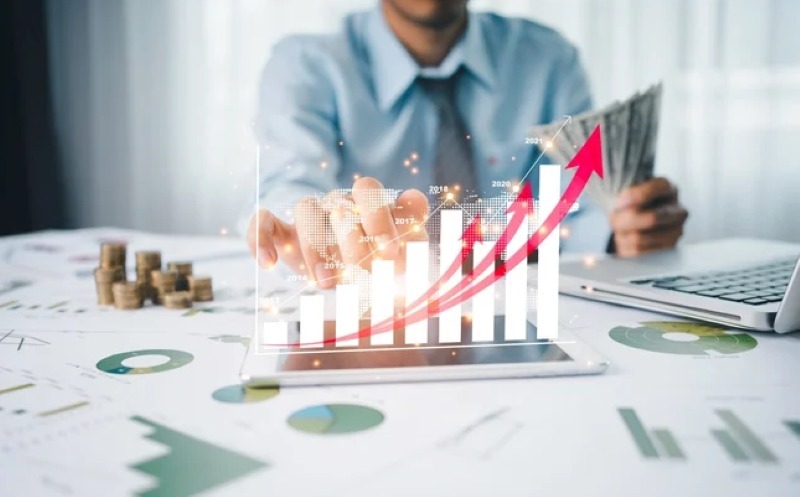 Người ngồi tại bàn làm việc, sử dụng máy tính bảng hiển thị biểu đồ tăng trưởng, bên cạnh là tiền mặt và tài liệu.