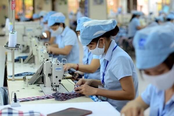 Công nhân trong xưởng may, đeo khẩu trang và đồng phục, tập trung làm việc trên máy may công nghiệp.