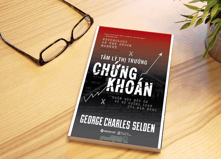 Cuốn sách về tâm lý thị trường chứng khoán trên bàn gỗ, bên cạnh là một cặp kính và cây xanh.
