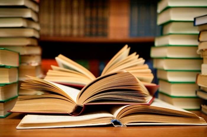 Những cuốn sách mở ra trên bàn, xung quanh là các chồng sách khác nhau trong một không gian thư viện.