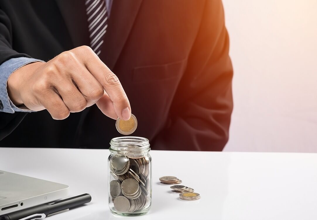 Người mặc vest đen bỏ đồng xu vào lọ thủy tinh chứa nhiều đồng xu khác trên bàn trắng.