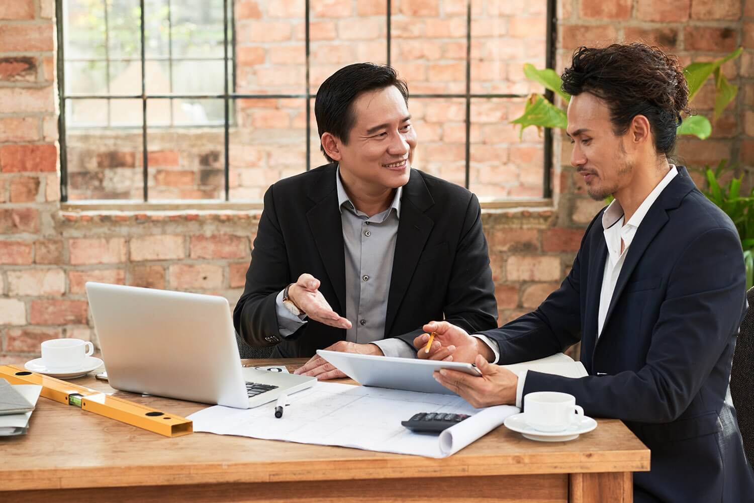 Hai người đàn ông ngồi tại bàn làm việc, thảo luận với máy tính xách tay và tài liệu.