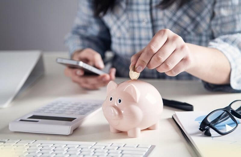 Người đang bỏ tiền xu vào lợn đất trên bàn làm việc, bên cạnh là máy tính và điện thoại.