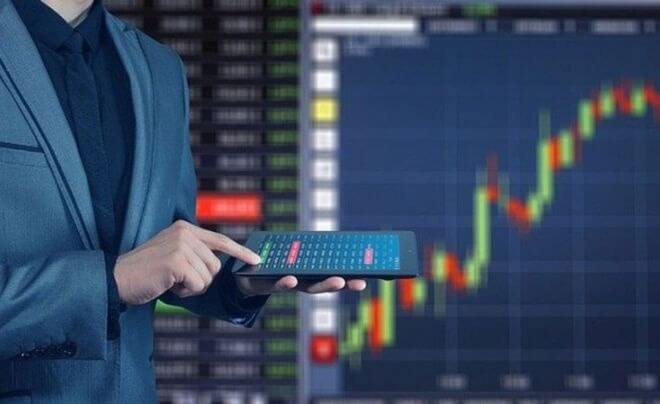 Người cầm máy tính bảng trước màn hình hiển thị biểu đồ chứng khoán với các cột và đường xu hướng.