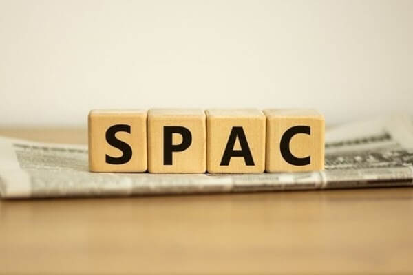 Các khối gỗ xếp thành chữ "SPAC" đặt trên một tờ báo, nền màu gỗ nhạt.