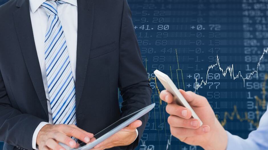 Người mặc vest cầm máy tính bảng, người khác cầm điện thoại, nền là biểu đồ tài chính với số liệu.