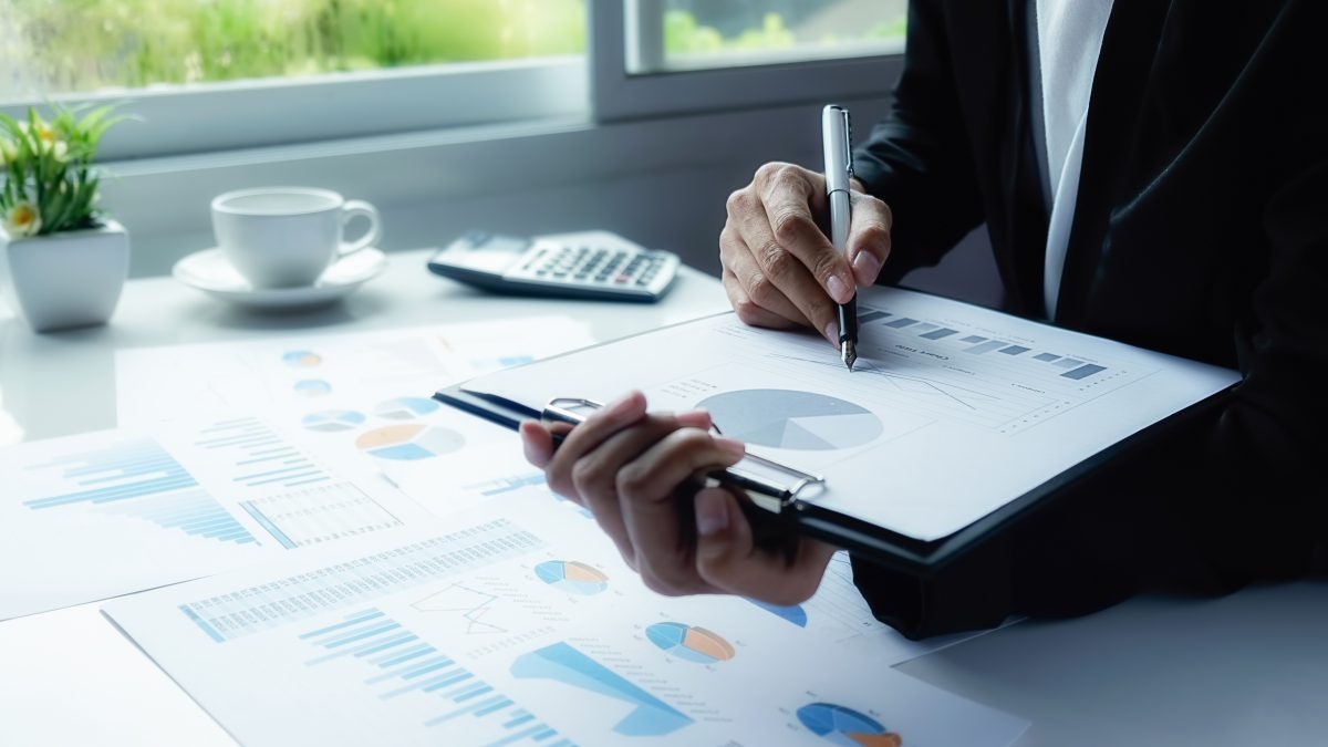 Người đang xem tài liệu biểu đồ và ghi chú, xung quanh có các tờ giấy, máy tính và cốc cà phê.
