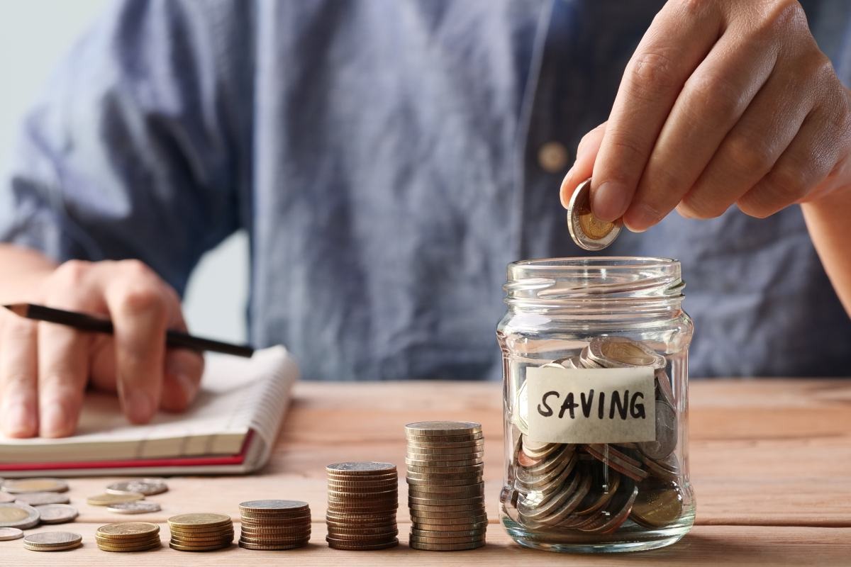 Người đang cho đồng xu vào lọ tiết kiệm, bên cạnh là sổ ghi chép và các chồng tiền xu.