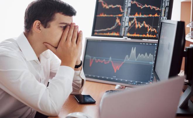 Người đàn ông ngồi trước màn hình máy tính với biểu đồ tài chính, vẻ mặt căng thẳng, tay che mặt.