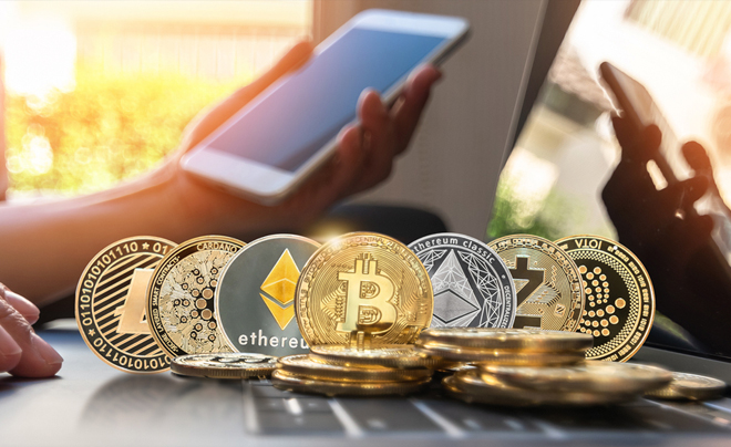 Người cầm điện thoại di động, phía trước là các đồng tiền mã hóa như Bitcoin, Ethereum trên bàn.