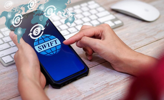 Người dùng đang thao tác trên điện thoại có logo SWIFT, bàn phím và chuột máy tính ở phía sau.