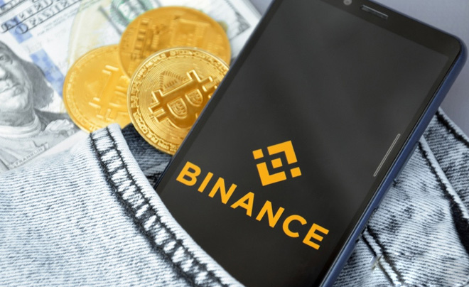 Điện thoại hiển thị logo Binance nằm trên quần jean, cạnh các đồng tiền bitcoin và tiền đô la Mỹ.