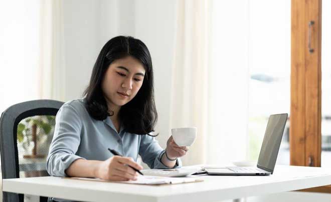 Người phụ nữ ngồi tại bàn làm việc, tay cầm bút và cốc, trước mặt là máy tính xách tay, ánh sáng tự nhiên.