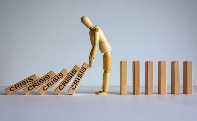 Một hình nhân gỗ đứng cạnh các khối chữ "Crisis" đang đổ, biểu tượng cho sự can thiệp vào khủng hoảng.