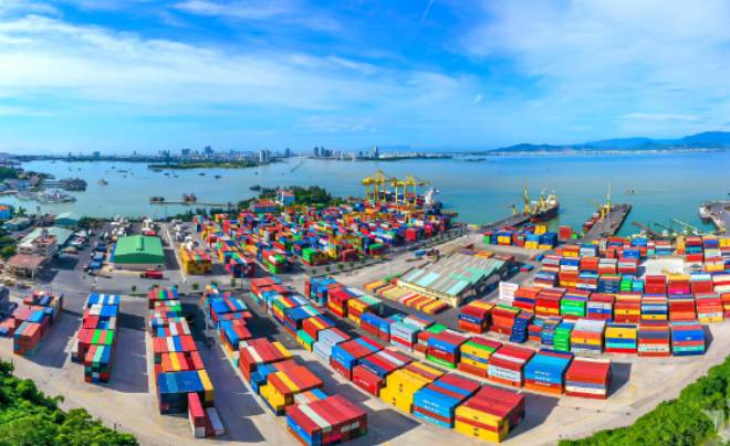Cảng biển với nhiều container màu sắc, cần cẩu và bến cảng, xa xa là thành phố và biển dưới bầu trời xanh.