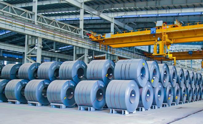Các cuộn thép lớn xếp chồng trong nhà kho công nghiệp, với cần cẩu vàng phía trên và kiến trúc trần cao.