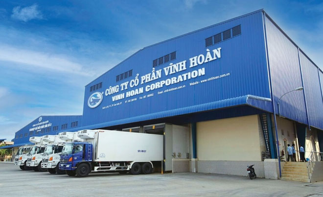 Tòa nhà công ty lớn màu xanh, phía trước có nhiều xe tải trắng đang đậu.