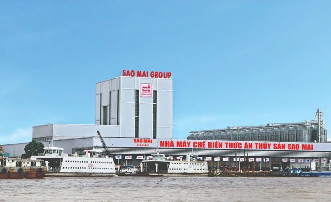 Nhà máy chế biến thức ăn thủy sản bên bờ sông, với tàu thuyền neo đậu phía trước, trời xanh và nắng nhẹ.