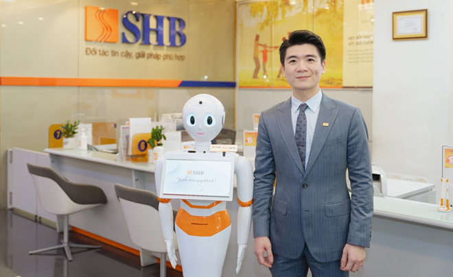 Người đàn ông trong bộ vest đứng cạnh robot trong sảnh ngân hàng, phía sau là bàn làm việc và logo SHB.