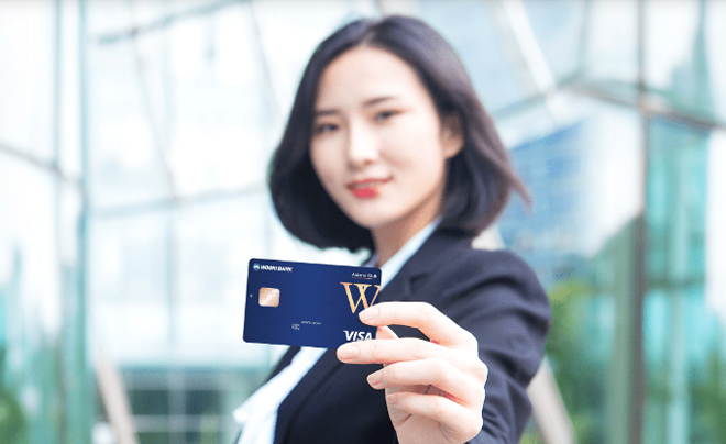 Người phụ nữ giơ thẻ tín dụng màu xanh dương, đứng trước phông nền mờ của các tòa nhà.