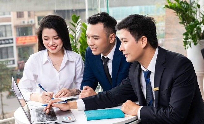 Ba người mặc vest và áo sơ mi ngồi quanh bàn, cùng nhau xem máy tính xách tay, có cây xanh bên cửa sổ.