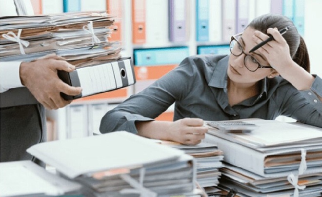 Người phụ nữ ngồi bàn làm việc với nhiều tài liệu, tỏ vẻ mệt mỏi khi nhìn người khác đang đưa thêm hồ sơ.