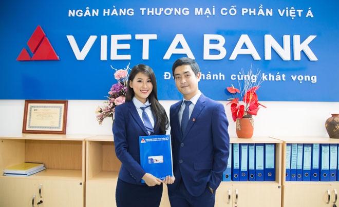 Hai người trong trang phục công sở đứng trước logo Viet A Bank, cầm tài liệu. Văn phòng trang trí hoa và tủ hồ sơ.