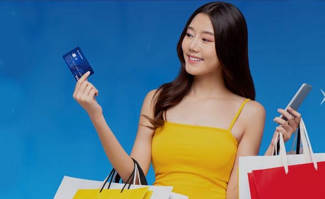 Người phụ nữ mặc áo vàng cầm thẻ ngân hàng, túi mua sắm và điện thoại, nền xanh.