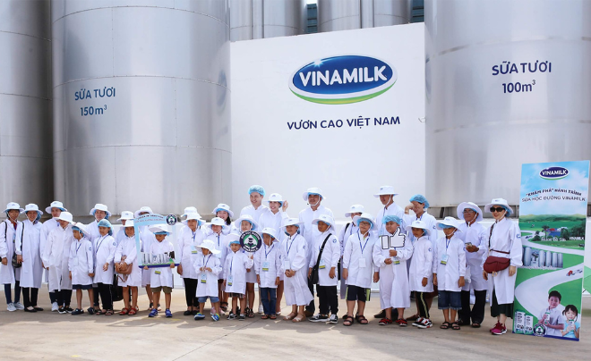 Nhóm người mặc áo bảo hộ trắng đứng trước nhà máy có logo Vinamilk, xung quanh là các bồn chứa lớn.