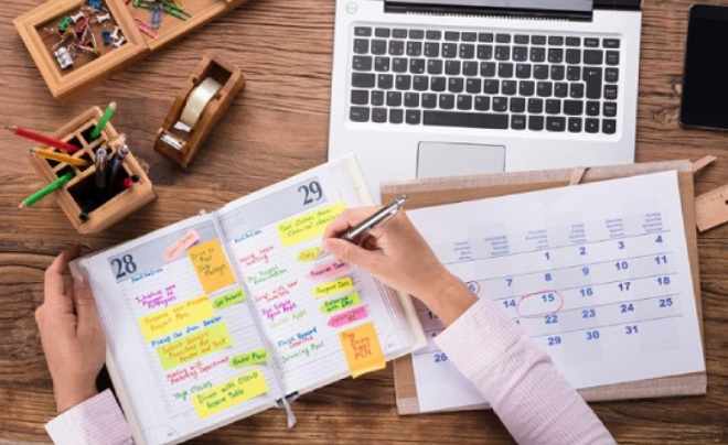 Người đang viết kế hoạch vào sổ tay, bên cạnh là máy tính xách tay và lịch tháng trên bàn gỗ.